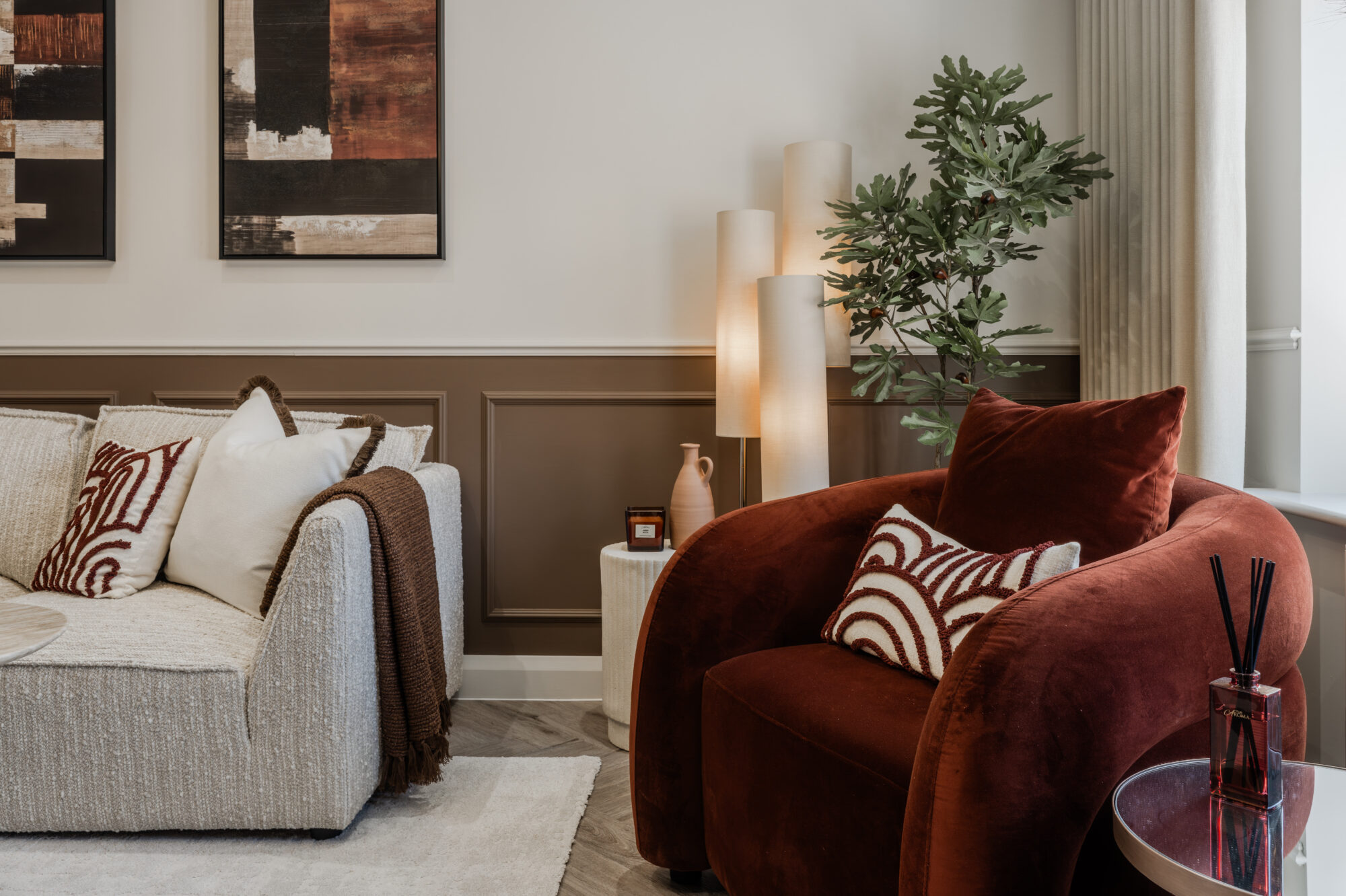 Living room with a cream boucle sofa and rust-coloured velvet armchair, styled with patterned cushions, a fig leaf plant, cylindrical lamps and abstract wall art.